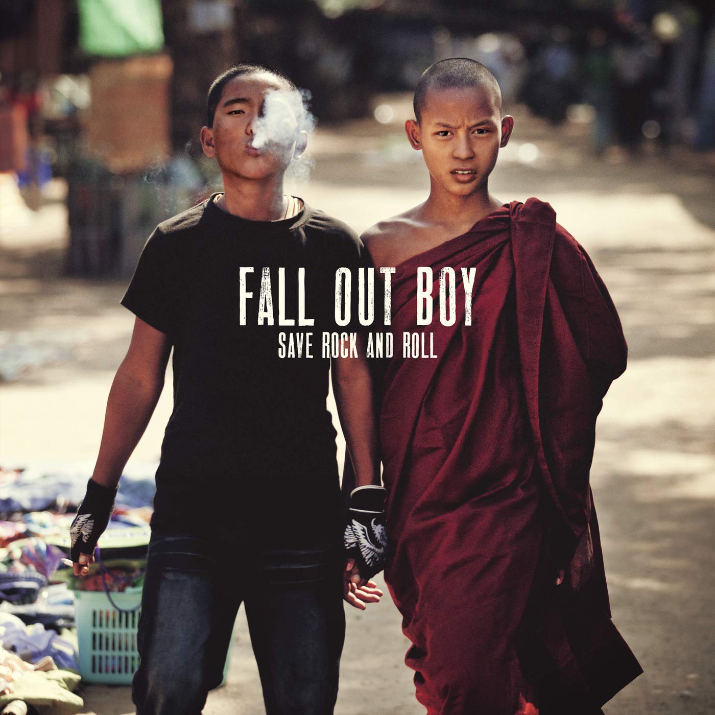 Cover for the album "Save Rock and Roll" by the pop-punk band Fall Out Boy. Depicted are two Asian boys holding hands, each with close-cropped hair. The boy on the left wears a black T-shirt, black fingerless gloves with white wings printed on them, and blue jeans. He exhales smoke at the viewer. The boy on the right wears red monk's robes tied over his left shoulder and stares at the viewer with a furrowed brow. Behind them is a street scene in broad daylight that includes a basket piled with objects and clothes laid out on the ground.