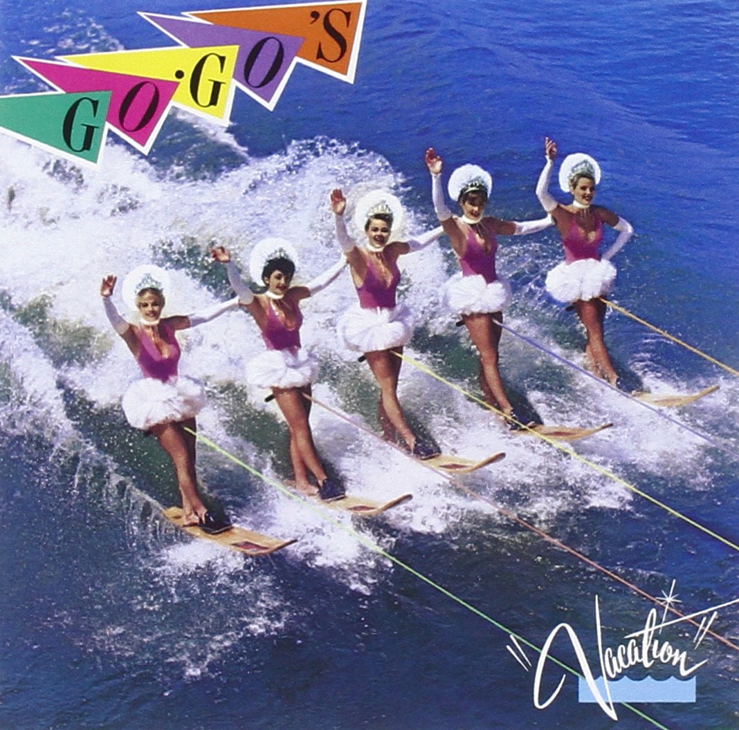 Cover for the album "Vacation" by the rock band The Go-Go's. Depicted are the five band members in a row on jet skis, their left arms placed on each other's shoulders while their right arms wave at the viewer. Each woman wears a tiara with white feathers in her hair, a pink leotard with white arm sleeves, and a white tutu. They are being pulled across ocean waves. The band's name is written with each letter on a different color triangular pennant in the top left corner of the frame. The bottom right corner of the frame displays the album name in white script with quotes over a cartoon graphic of ocean waves.